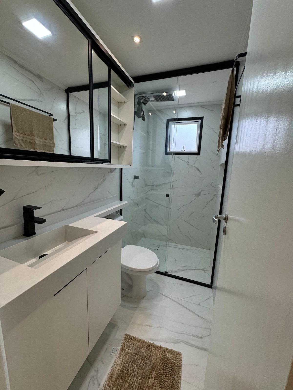 A modern bathroom features a walk-in shower enclosed by glass. A white countertop with a sink is set against marble-patterned walls. Neutral tones create a clean and spacious look, while natural light filters in from a small window.