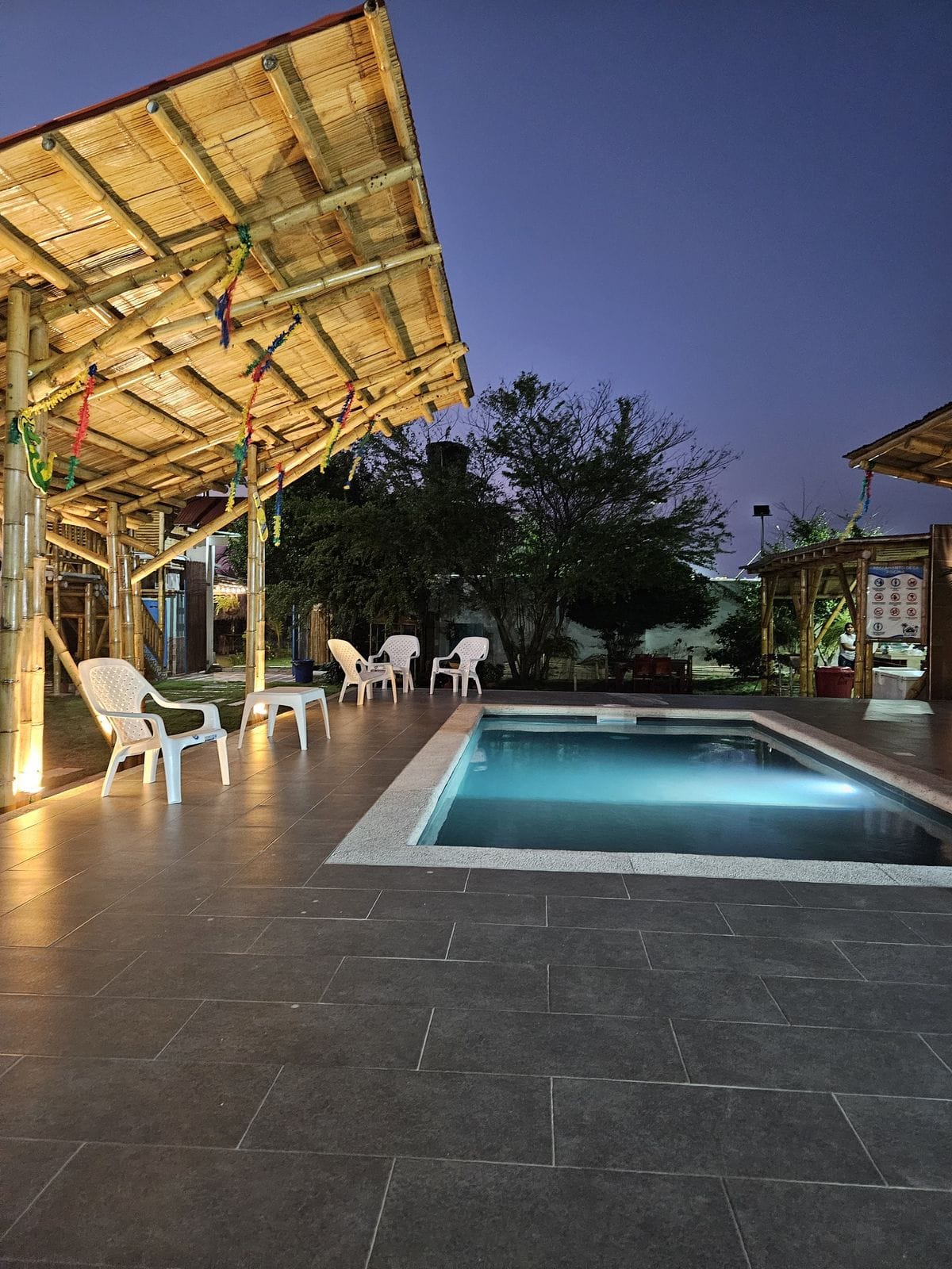 A private pool area is illuminated by soft lighting, with a rectangular pool surrounded by a tiled deck. Several white chairs are positioned around the space, and open, thatched-roof structures provide shade, enhancing the relaxed outdoor setting.