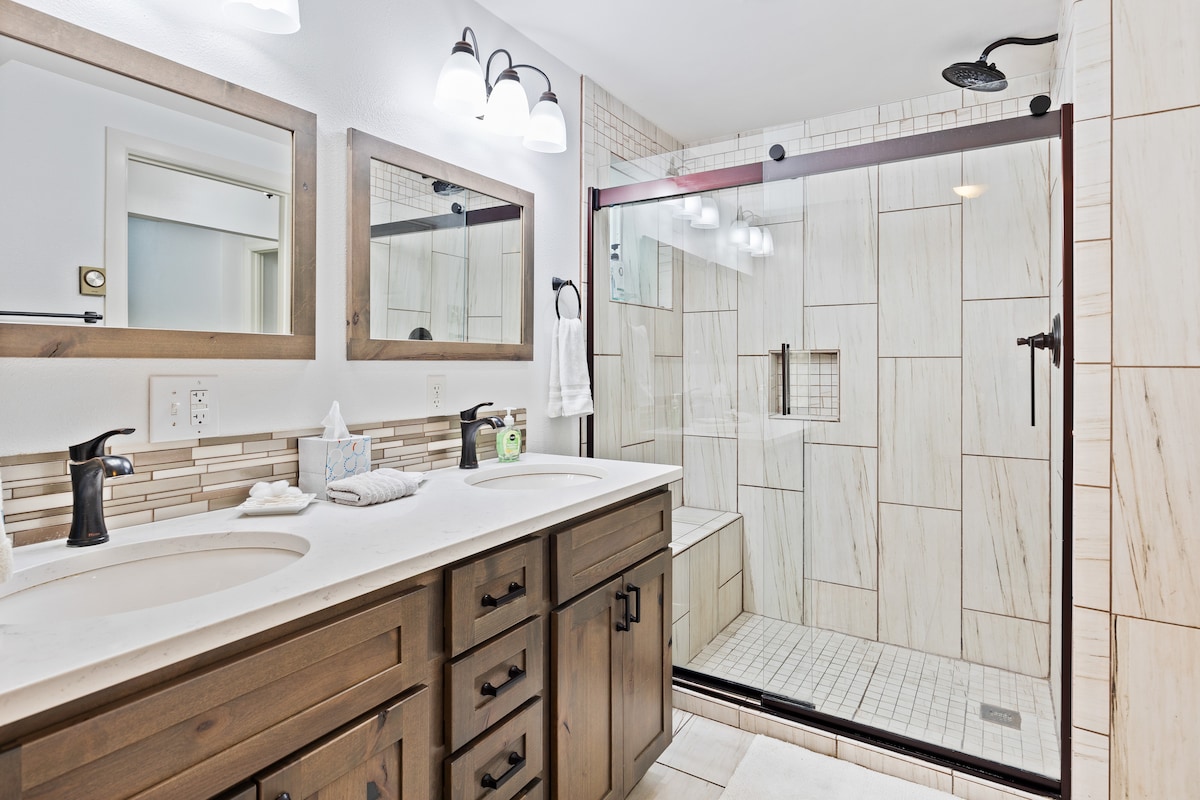 The bathroom features a double sink vanity with dark wooden cabinetry and modern fixtures. A spacious walk-in shower is lined with light-colored tiles and includes built-in shelving. Two mirrors are positioned above the sinks, providing ample light from overhead fixtures.