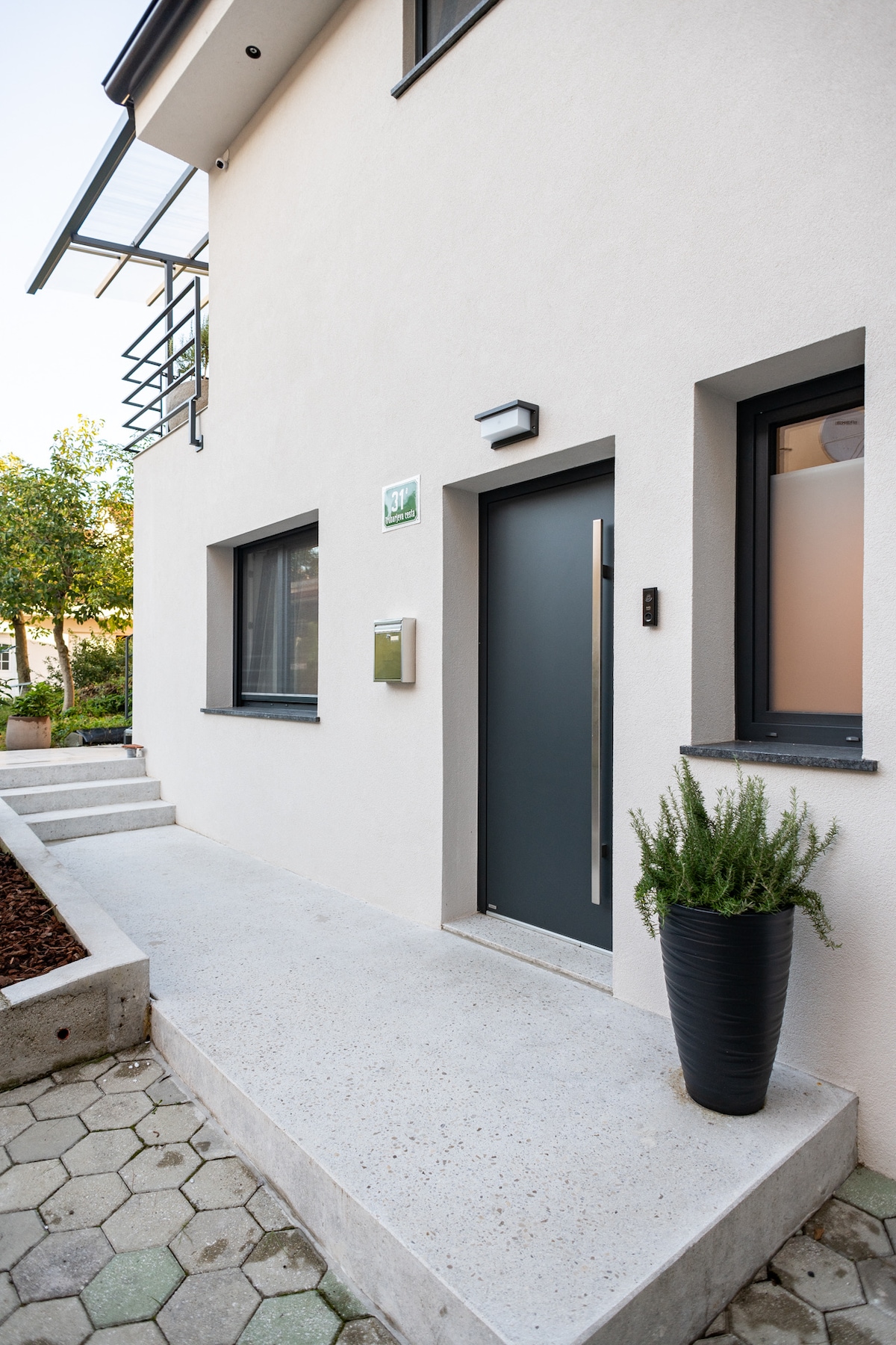 The exterior entrance is framed by a neatly paved pathway leading to a modern door with a sleek handle. A tall planter with greenery adds a touch of nature, while large windows provide ample light. The building features light-colored walls and a minimalist design.