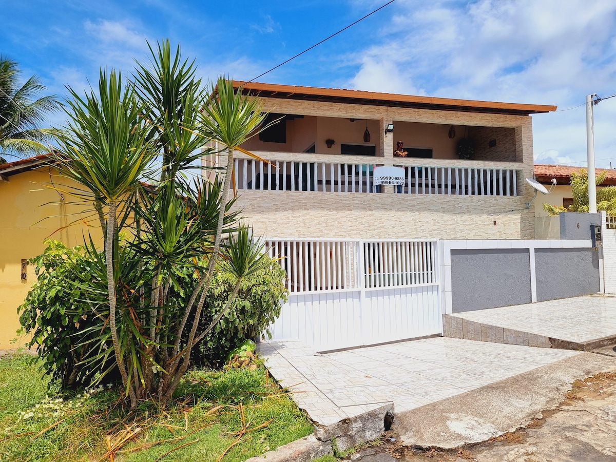 A two-story house is displayed with a welcoming exterior. The entrance is framed by landscaped greenery and palm plants, while the white gate provides access to the property. A bright blue sky is featured above the structure, emphasizing the outdoor space.