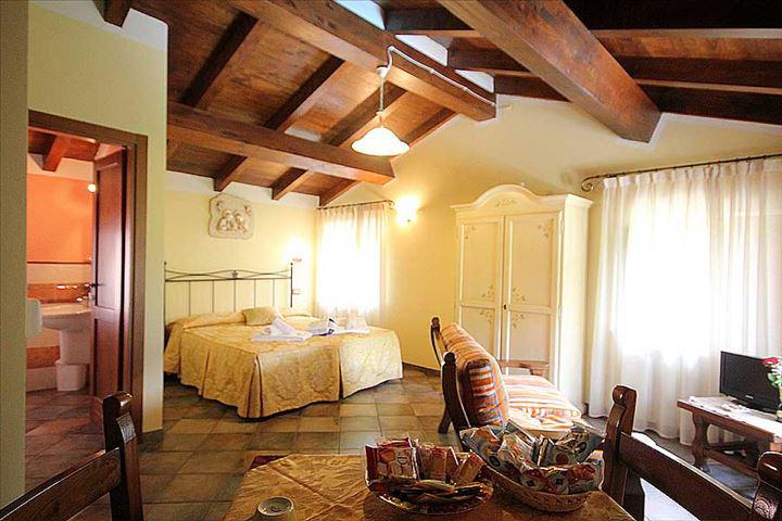 An open space studio features a warm color palette with exposed wooden beams. A seating area with a couch is visible, alongside a spacious bed covered in a light yellow bedspread. A wooden dining table is set with snacks, while soft natural light filters through sheer curtains.