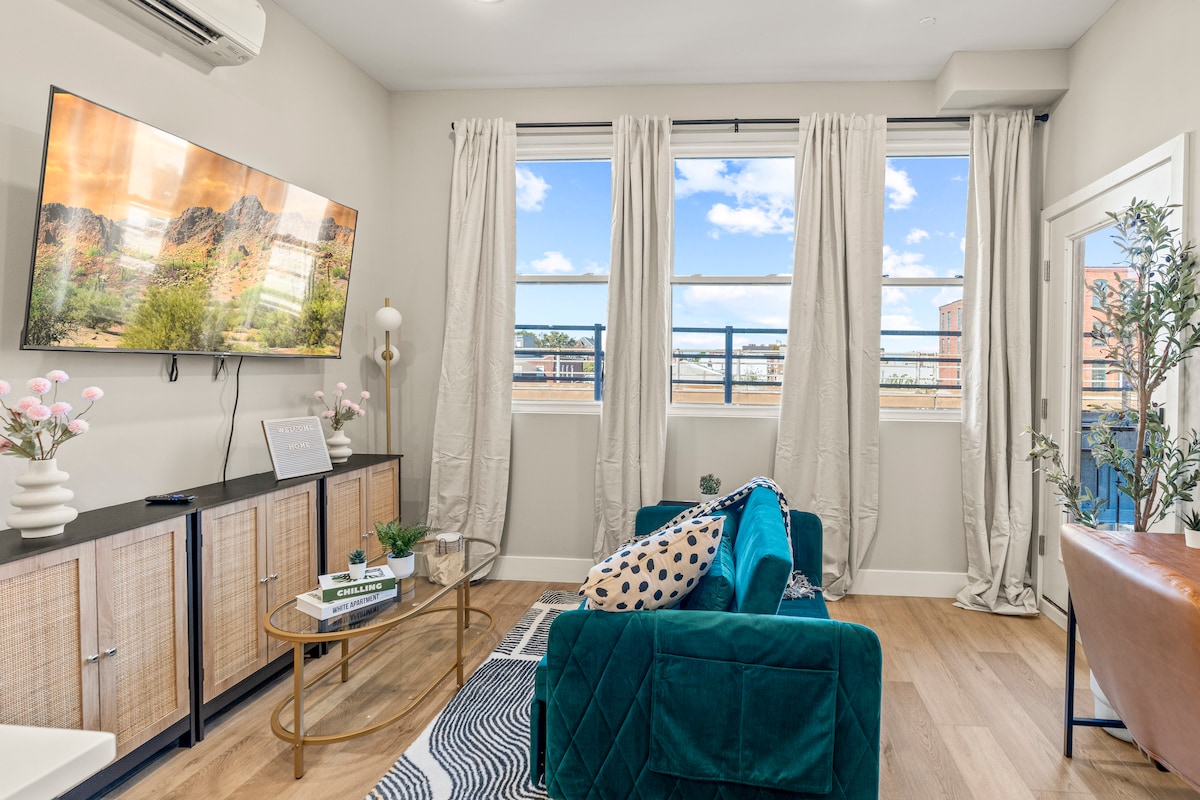The open-concept living area features large windows allowing natural light to fill the space. A teal couch is positioned in front of a media console, while a round coffee table sits nearby. Light-colored curtains frame the windows, enhancing the inviting atmosphere.