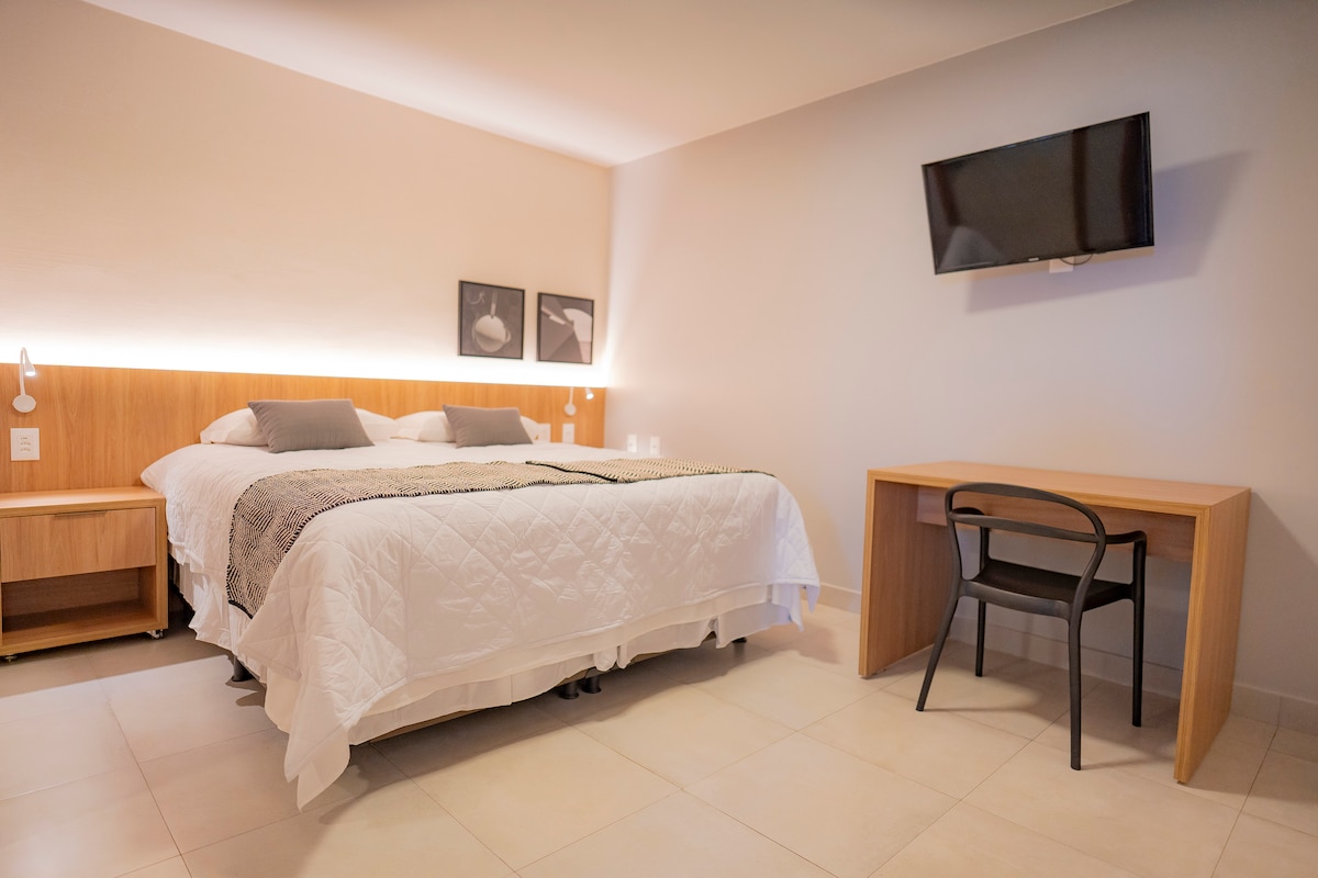 A spacious sleeping area features a king size bed with neutral bedding and decorative pillows. A television is mounted on the wall opposite a wooden desk with a chair. Soft lighting highlights the modern decor, complemented by framed artwork.