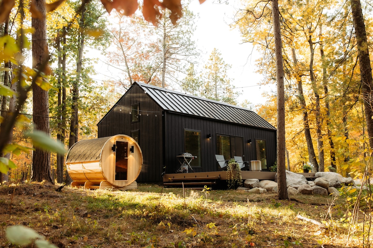 The exterior of a modern cabin is set amid tall trees, showcasing a sleek black design. A cedar barrel sauna is positioned on the deck next to smooth rocks. Sunlight filters through autumn leaves, creating a warm, inviting environment.