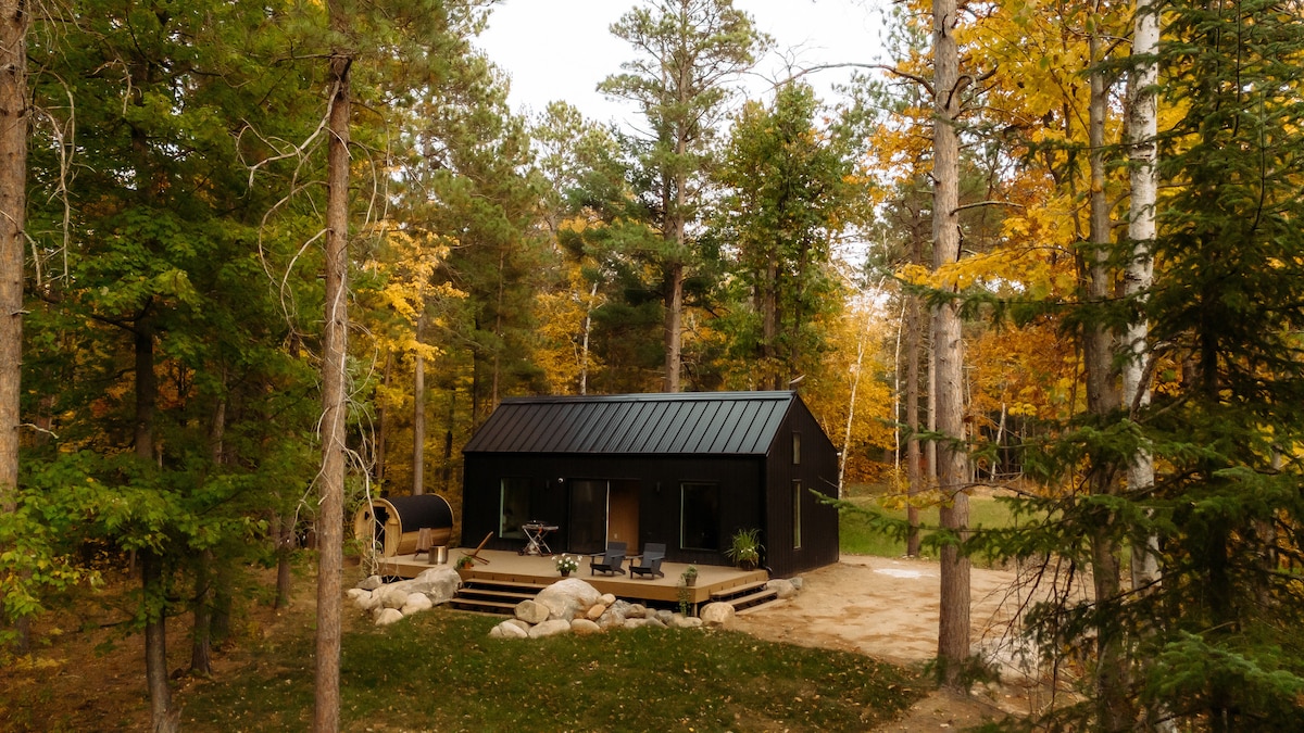 A modern black cabin is set among autumn-colored trees in a forested area. A spacious deck with seating arrangements and a cylindrical cedar sauna is visible. Natural stone steps lead up to the entrance, blending harmoniously with the surrounding landscape.