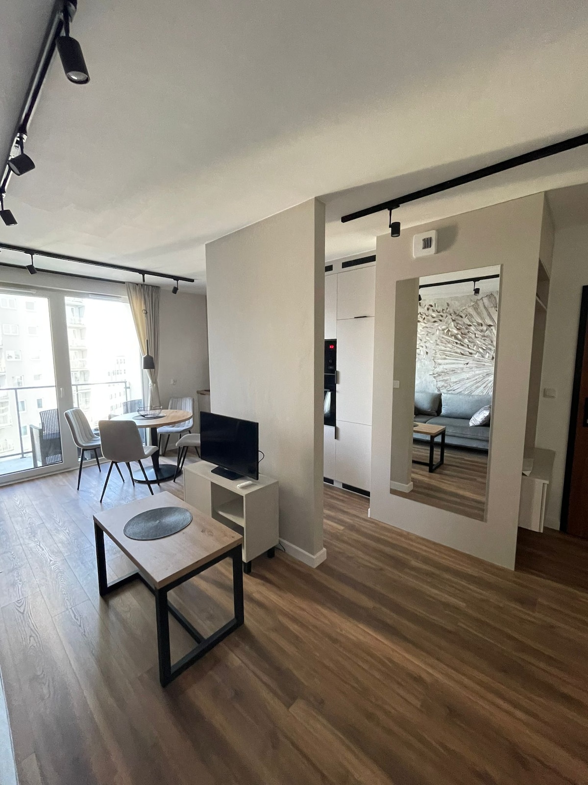 The living area is featured with a wooden floor and a cozy seating arrangement. A minimalist coffee table sits in front of a small television. A dining set for four is positioned near the window, allowing natural light to fill the space. A mirror reflects the adjacent bedroom.