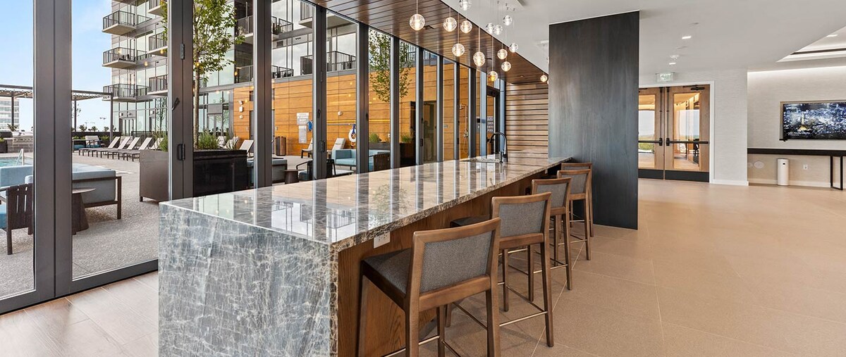 A spacious lounge area features a modern granite countertop with several wooden bar stools. Large windows provide a view of the outdoor pool area, allowing natural light to enhance the stylish decor. Pendant lighting hangs above, adding a sophisticated touch to the ambiance.