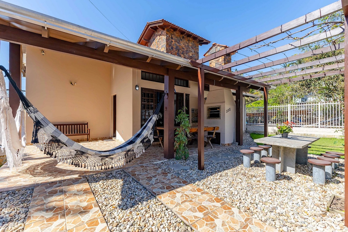 The outdoor patio features a stone floor and is shaded by a wooden pergola. A hammock is suspended for relaxation, while a large stone table and several benches provide ample seating for gatherings. Lush greenery is visible in the background, enhancing the tranquil environment.