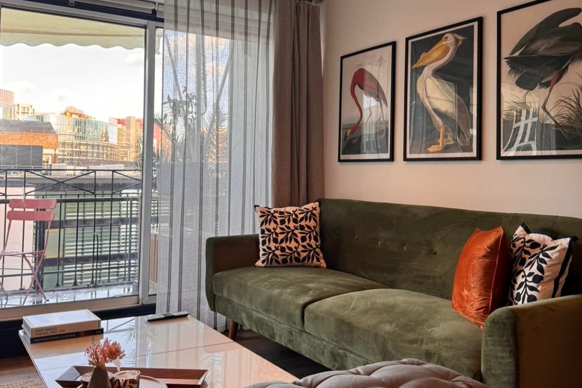 A living area showcases a plush green sofa adorned with decorative pillows, positioned near floor-to-ceiling sliding glass doors. Bright sunlight filters through sheer curtains, illuminating framed artwork on the wall. A small balcony can be glimpsed beyond, offering a view of the outdoors.