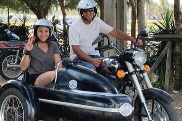 A classic motorcycle with a sidecar is parked in a shaded outdoor area. Two riders are wearing helmets, with one seated in the sidecar and the other on the bike. Lush greenery and other vehicles are visible in the background.