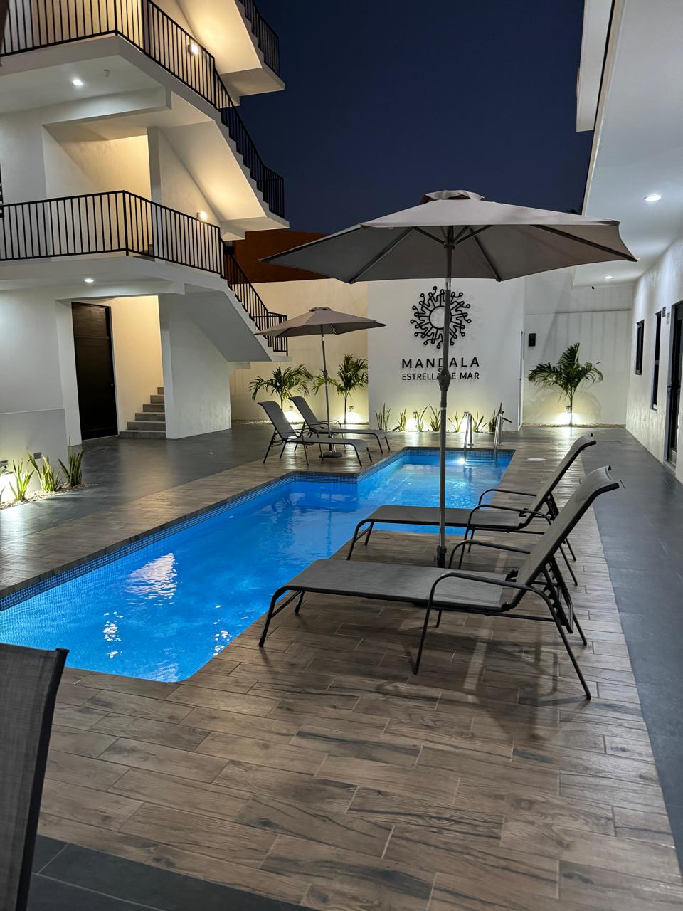 A tranquil pool area is featured, enhanced by two lounge chairs and a large umbrella for shade. Soft lighting illuminates the pool, reflecting off the water's surface. The surrounding space includes plants in planters and a modern building facade in the background.