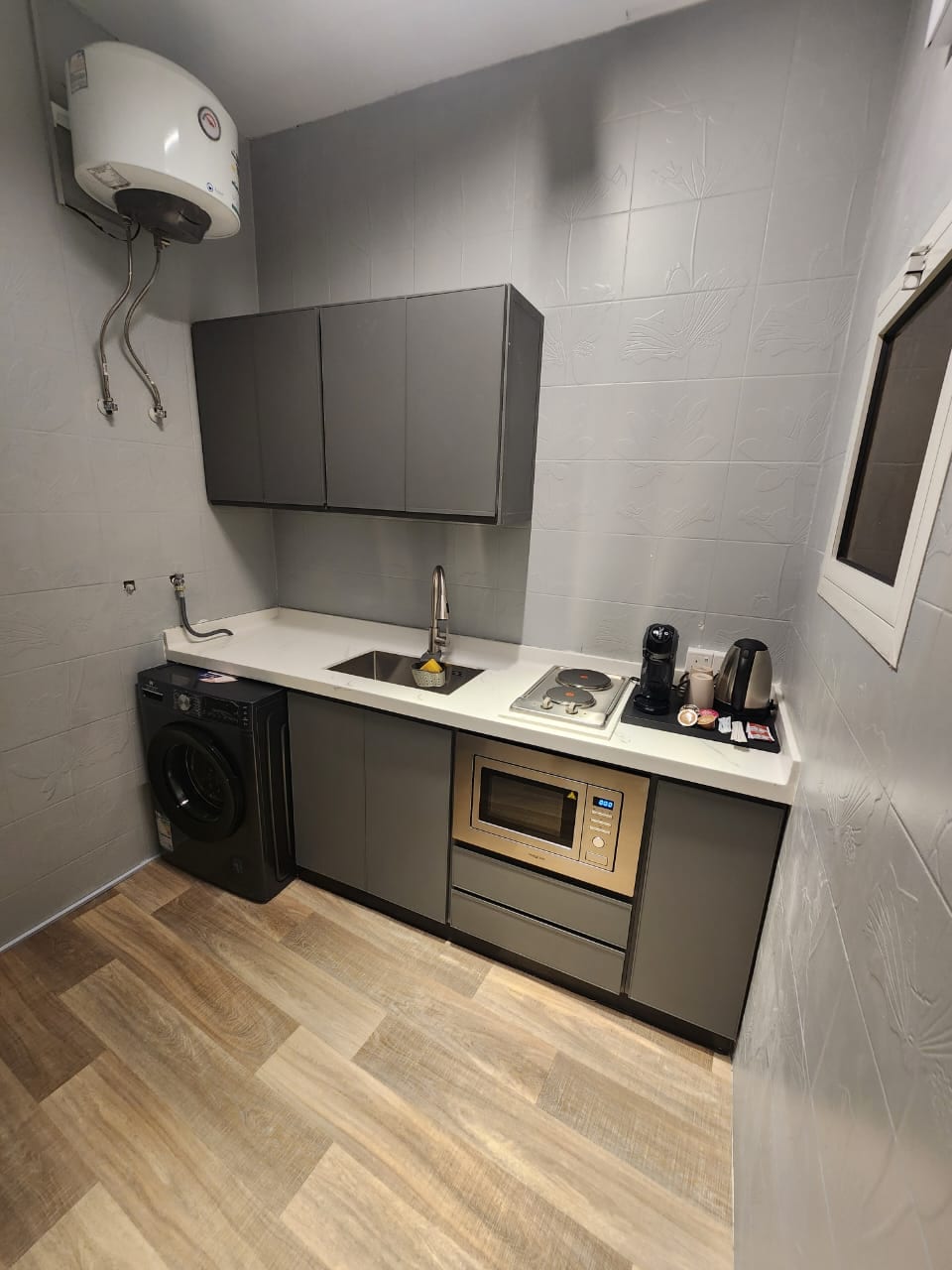 The kitchen area is equipped with modern gray cabinetry and a white countertop. A sink and an electric stove are integrated into the design, alongside a washing machine and a microwave. Electrical appliances, including a coffee maker, are present, offering convenience for guests.