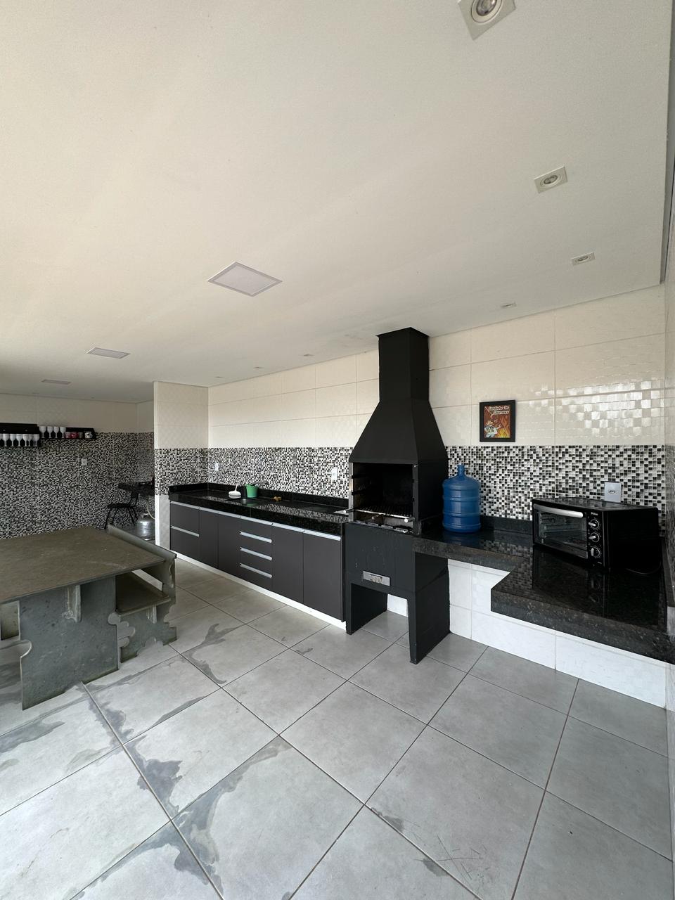 A modern kitchen area features sleek black cabinets and a dark granite countertop. An outdoor grill is positioned nearby, alongside a microwave and a large water dispenser. The tiled floor extends throughout the space, enhancing its spacious feel.