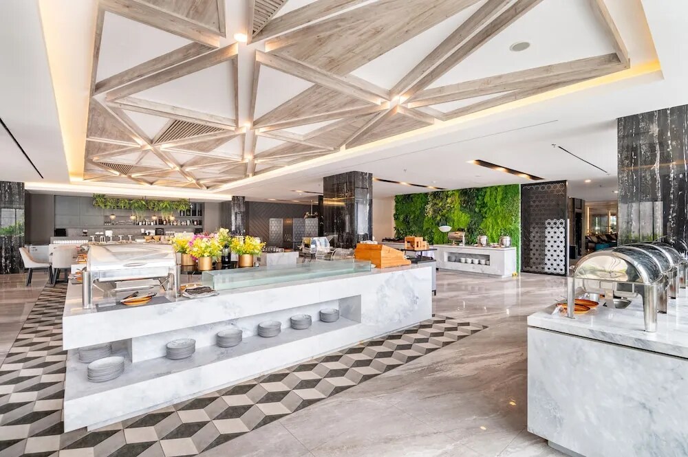 A modern dining area is highlighted by a large buffet counter made of marble, featuring various culinary displays. Elegant geometric patterns adorn the floor, complemented by lush greenery on the walls. Natural light filters through, enhancing the welcoming ambiance of the space.