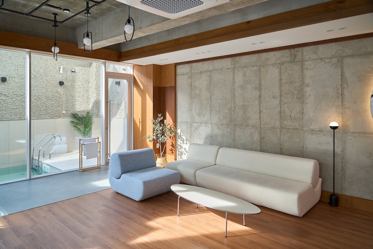 A spacious living area features minimalistic furniture arrangements, including a light-colored sofa and unique armchair. Large glass doors offer views of a private outdoor pool area, while natural wood accents and neutral tones create a calming environment.