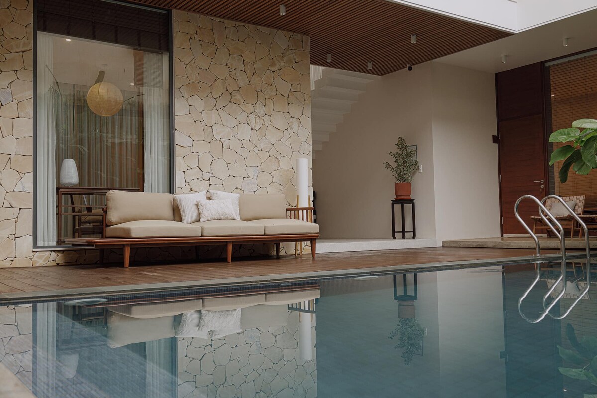 A serene outdoor space features a wooden deck surrounding a tranquil swimming pool. A modern sofa with neutral-toned cushions is positioned near the water, while a small potted plant and a stylish side table add subtle greenery. Light reflects gently off the pool's surface.