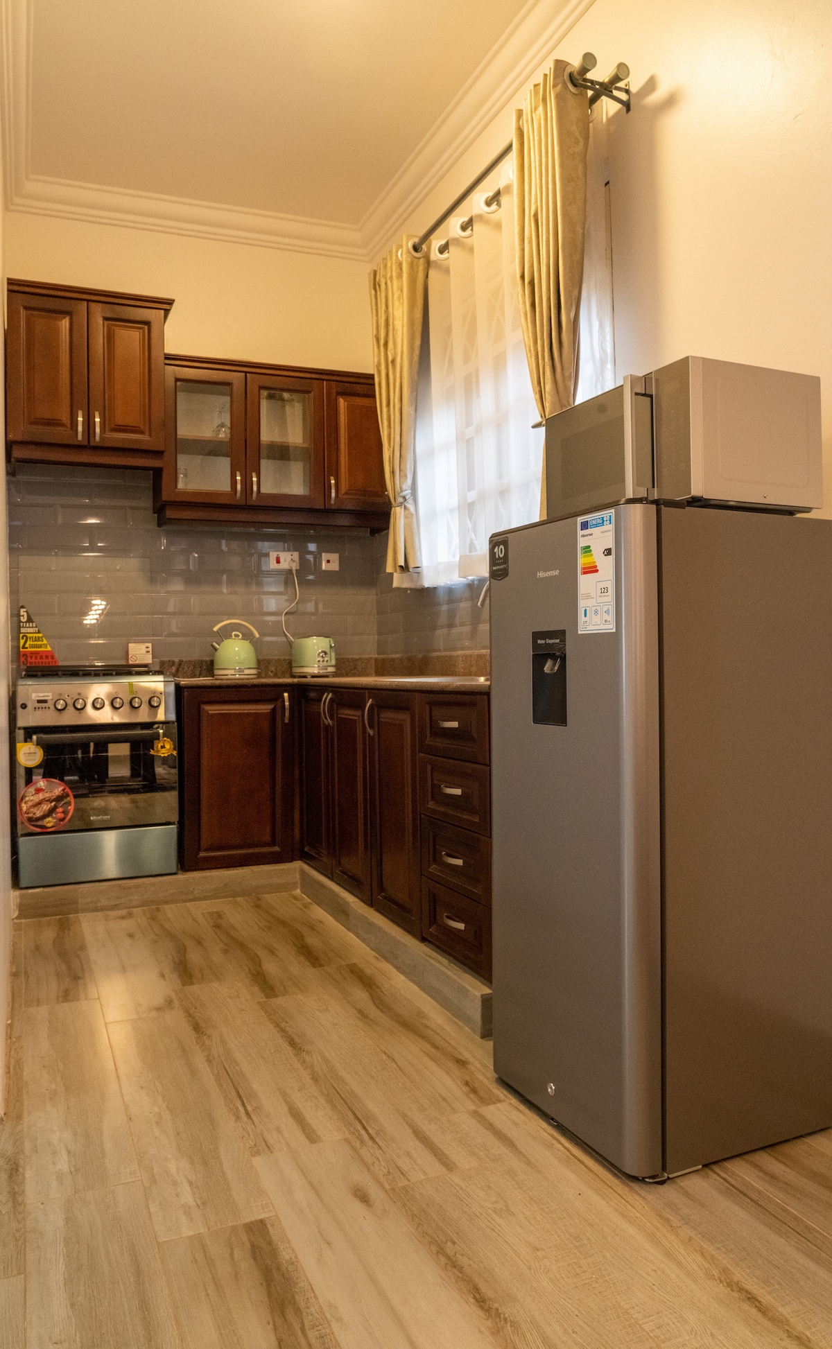 The kitchen features wooden cabinets and modern appliances, including a refrigerator and microwave. A gas stove is visible, complemented by a tiled backsplash. Natural light enters through the window, and essential cooking items are easily accessible, contributing to a functional cooking space.