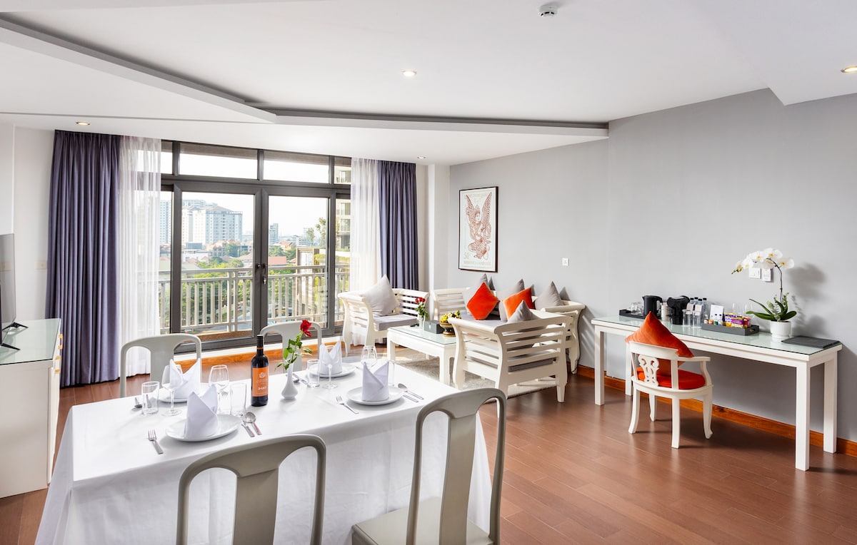 A bright and airy dining area features a neat dining table set for four, complemented by decorative tableware. Beyond, a comfortable seating arrangement is positioned beside large windows offering views of the city skyline. Light flows in, illuminating the warm wooden floor.