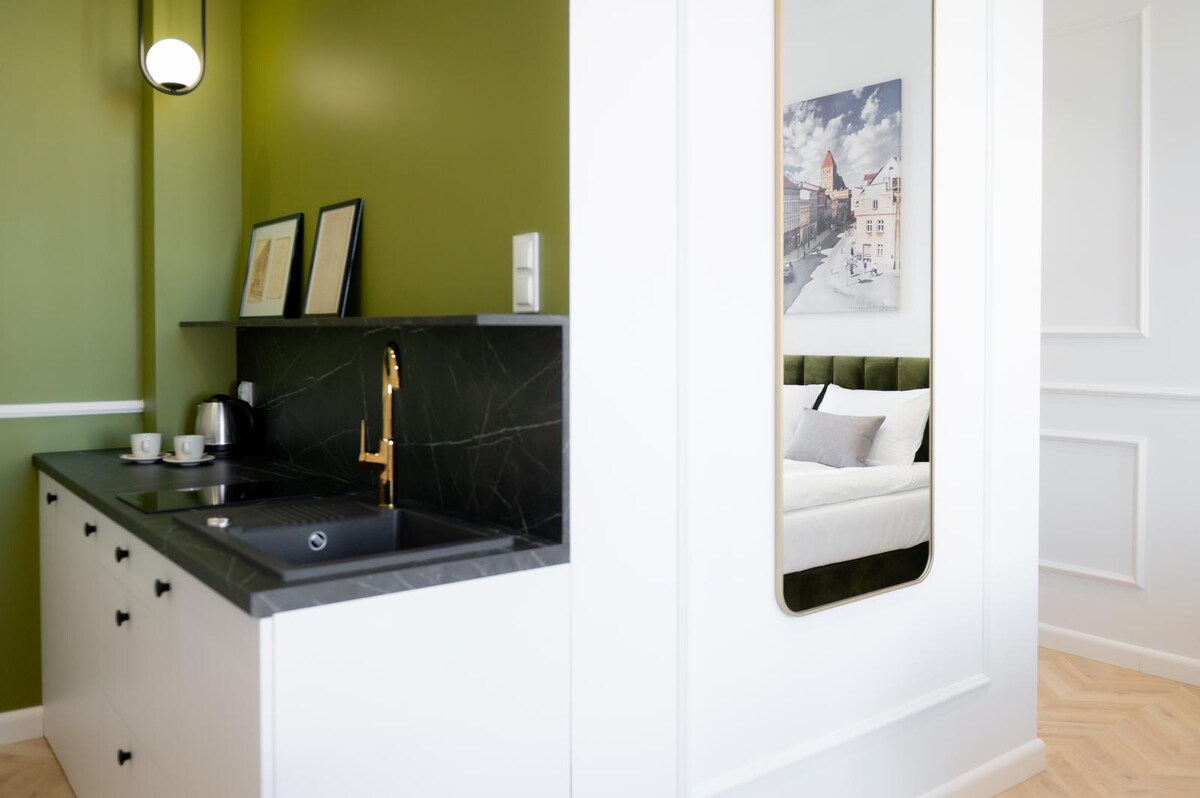 A modern kitchen area features dark cabinetry paired with a sleek black countertop. Gold fixtures add a touch of elegance. A green accent wall enhances the space's vibrancy. A mirror reflects the adjacent sleeping area, which is designed with neutral bedding and muted decor.