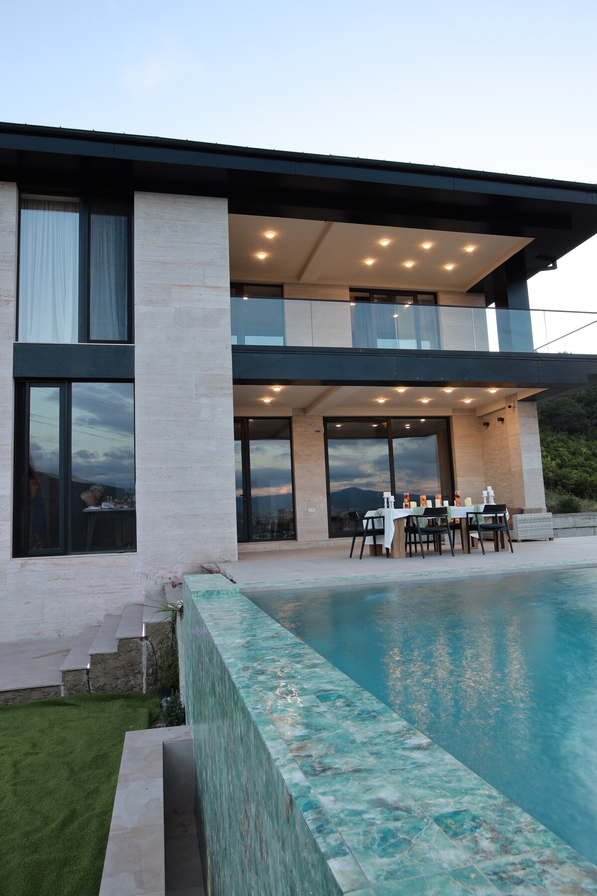 The villa exterior features a modern design with expansive glass windows reflecting the sky. A spacious patio overlooks a shimmering pool, where a dining table is set for guests. Lush greenery surrounds the area, enhancing the overall serene atmosphere.