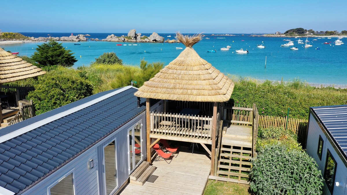 An elevated thatched terrace overlooks calm waters dotted with boats. The backyard features wooden decking and lounge chairs, framed by lush greenery and coastal scenery, creating a serene outdoor space that invites relaxation.