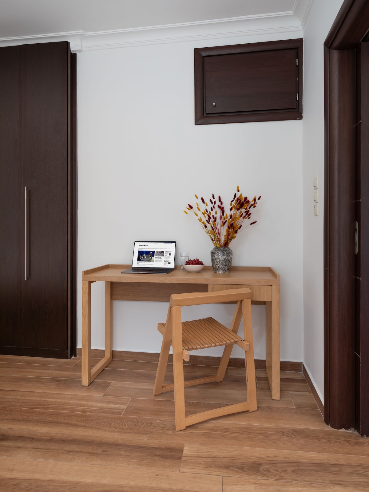 A compact workspace is featured, consisting of a light wood desk accompanied by a matching chair. A laptop sits on the desk, and a vase with colorful dried flowers adds a touch of decor. The warm wood flooring complements the modern design.