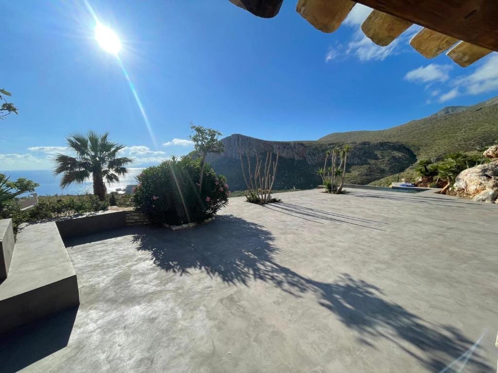 A spacious outdoor terrace is observed, featuring a smooth concrete surface. Lush greenery, including palm trees and flowering plants, is present, complemented by scenic mountain views in the background. Bright sunlight enhances the natural beauty of the surroundings, creating a serene outdoor space.