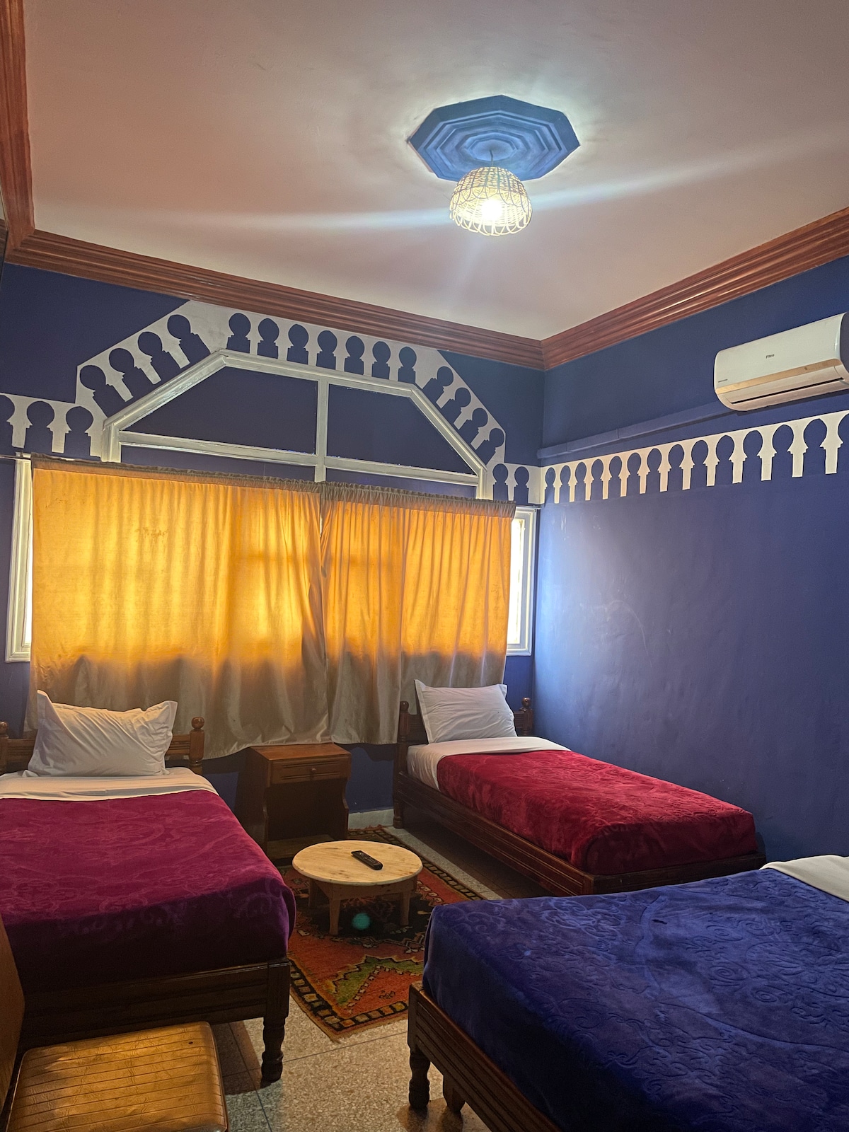 A cozy room features two twin beds covered in purple blankets and a wooden nightstand. Natural light streams in through large windows with golden curtains, creating a warm ambiance. Decorative wall patterns and a unique ceiling design are visible, while an air conditioning unit is mounted above.