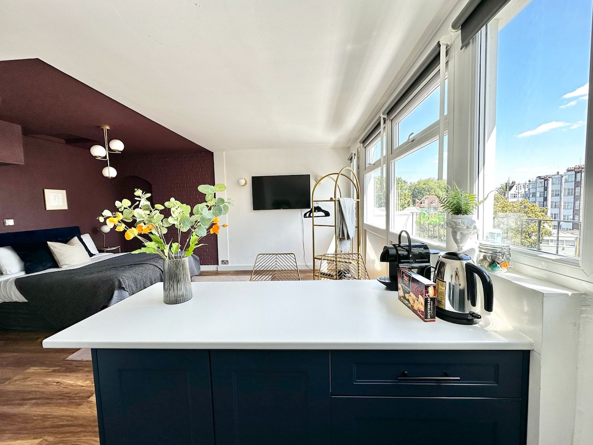 A view of the studio apartment showcases a modern kitchen with a dark blue countertop and a kettle. The seating area features a flat-screen TV and a stylish gold display rack. Large windows allow natural light to fill the space, enhancing the warm atmosphere.