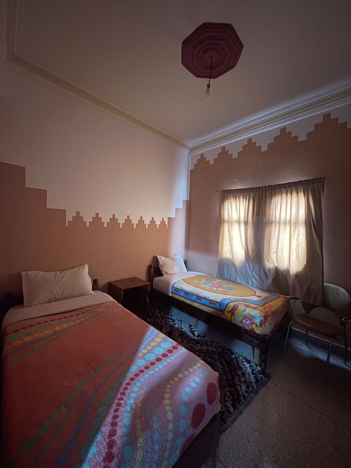 A modest guest room features two single beds dressed in colorful patterned blankets. A window allows natural light to filter in, complemented by light curtains. A small bedside table is positioned between the beds, and a simple chair is visible to the side, enhancing the space's functionality.