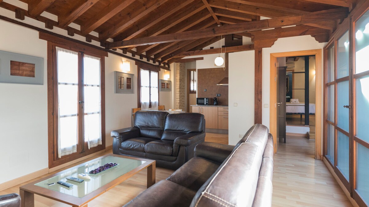 The spacious living area features wooden beams overhead and a combination of natural light and warm tones. Two dark leather sofas are positioned around a coffee table, with a kitchenette visible in the background. Glass doors lead to other areas of the apartment.