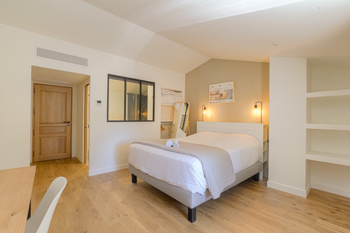 A well-lit bedroom features a spacious bed with a neutral-toned bedspread, complemented by wooden flooring. A stylish desk is positioned nearby, and a large mirror reflects natural light. Shelves are integrated into the design, and a door leads to another room, enhancing the area’s openness.