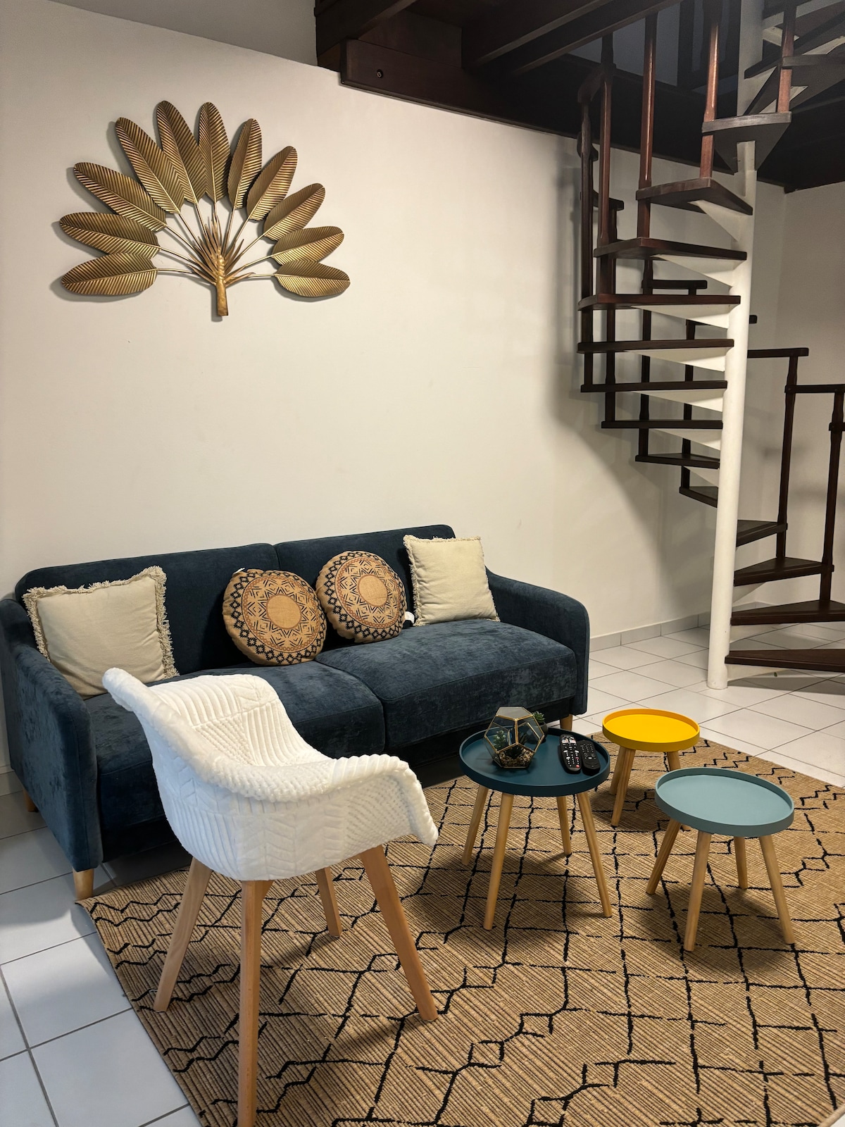 A cozy living area features a dark blue sofa adorned with decorative pillows, complemented by a textured area rug. A spiral staircase ascends in the background, while two small tables in yellow and teal offer a colorful contrast to the neutral tones of the room.