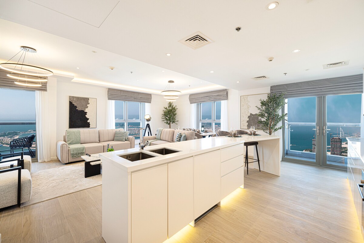 A spacious living area features modern furniture, including two sofas and armchairs beside large windows offering views of the coastline. A sleek kitchen island with integrated appliances complements the light wood flooring and neutral decor, creating an inviting atmosphere in the open layout.