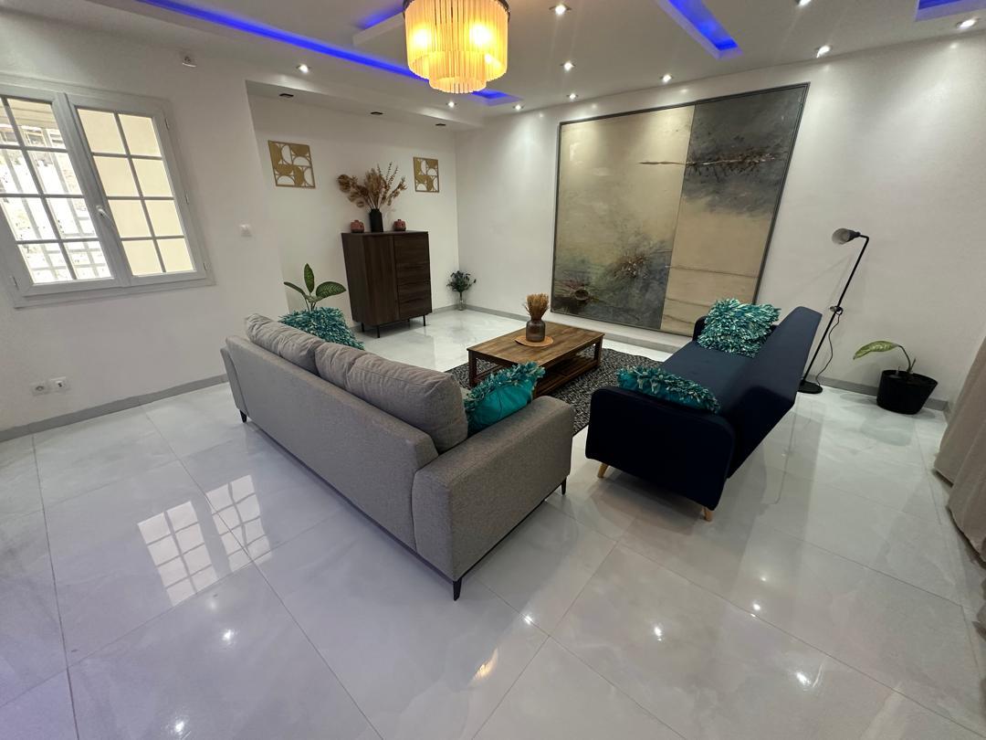 An inviting living area features two stylish sofas with decorative cushions, positioned on a glossy tile floor. A central coffee table sits atop a textured rug, with a large abstract artwork adorning the wall. Natural light enters through a window, enhancing the modern decor.