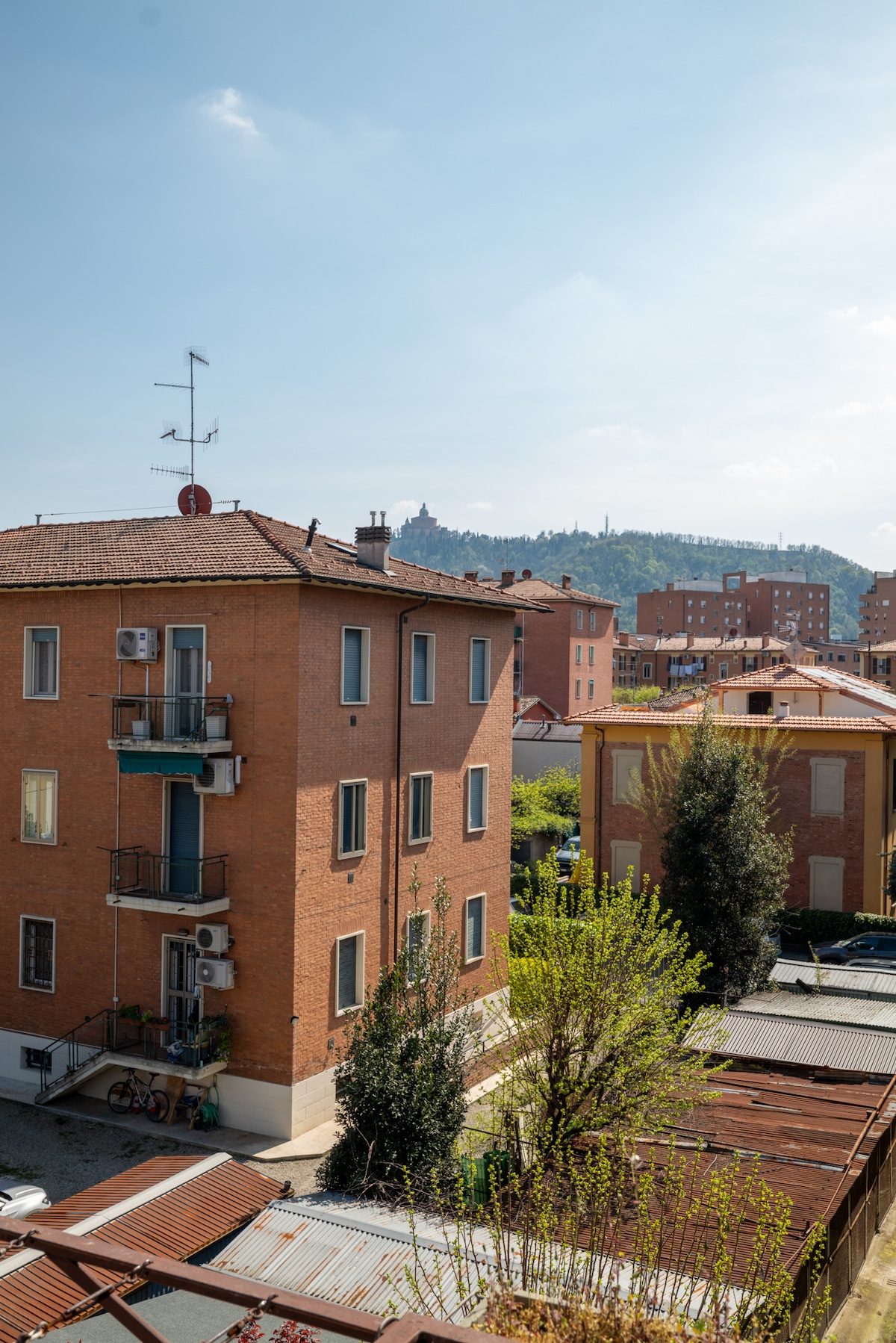 The image captures a view of a brick building with multiple windows and a balcony, set against a backdrop of rolling hills. Nearby structures and greenery offer a sense of tranquility, while the bright sky adds to the overall ambiance of the area.