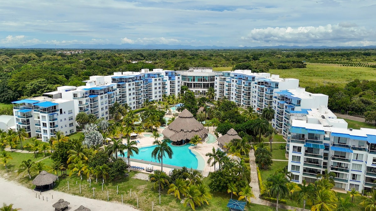 An aerial view showcases a modern complex surrounded by lush greenery and palm trees. A central pool is framed by tropical landscaping and a thatched-roof pavilion. Multiple buildings with balconies can be seen, inviting relaxation and enjoyment of the serene environment.