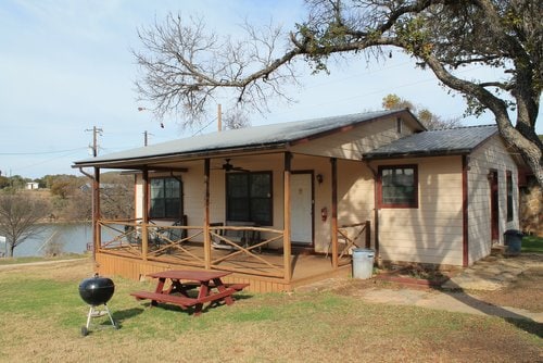 A two-bedroom cabin is shown with a spacious front porch featuring a picnic table and a charcoal grill. The exterior is complemented by light-colored walls and dark trim, surrounded by grass and trees. The lake is visible in the background.