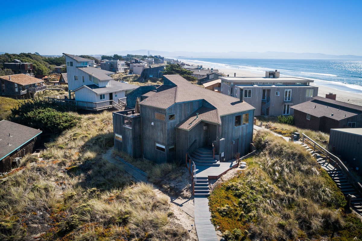 An aerial view of a contemporary beachfront house is captured, showcasing distinctive architectural features against a backdrop of sandy dunes and the ocean. Nearby homes are visible along the coastline, with clear skies and sunlight illuminating the scene.