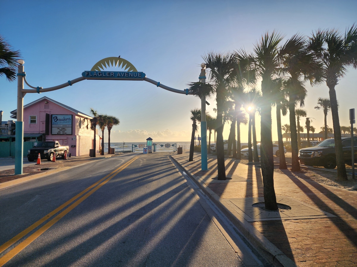 A welcoming entrance to Flagler Avenue is framed by tall palm trees. The golden light of dawn reflects off the ocean in the background, creating a serene atmosphere. A beachside establishment is visible to the left, accompanied by a clear path leading towards the water.