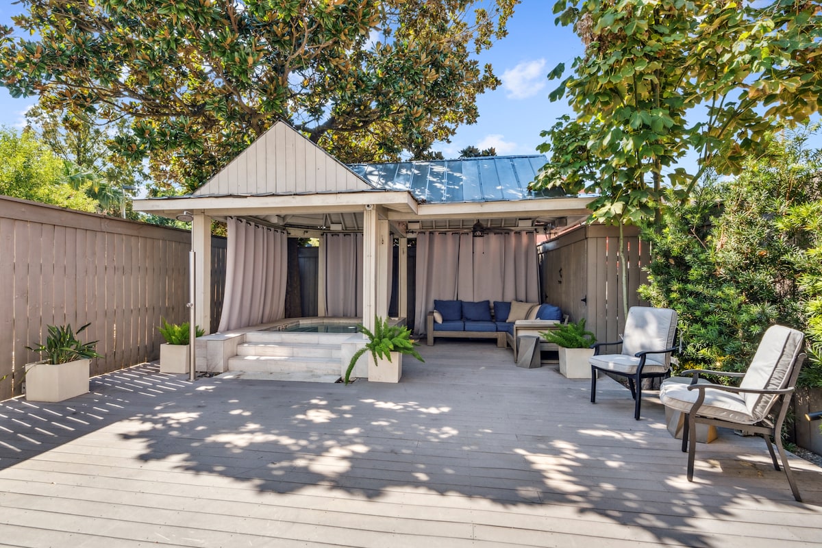 An outdoor patio area features a covered seating space with comfortable furniture. A hot tub is positioned nearby, surrounded by greenery and planters. The wooden deck is illuminated by natural light filtering through the trees, creating an inviting atmosphere for relaxation.
