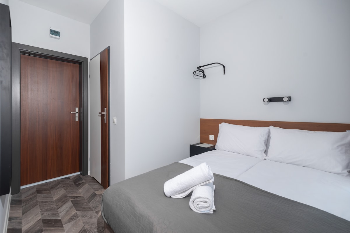 A well-lit sleeping area is present, featuring a spacious bed with crisp white linens. Neatly folded towels are arranged at the foot of the bed. A modern wall-mounted lamp provides additional lighting, while a wooden door is visible, leading to the hallway.