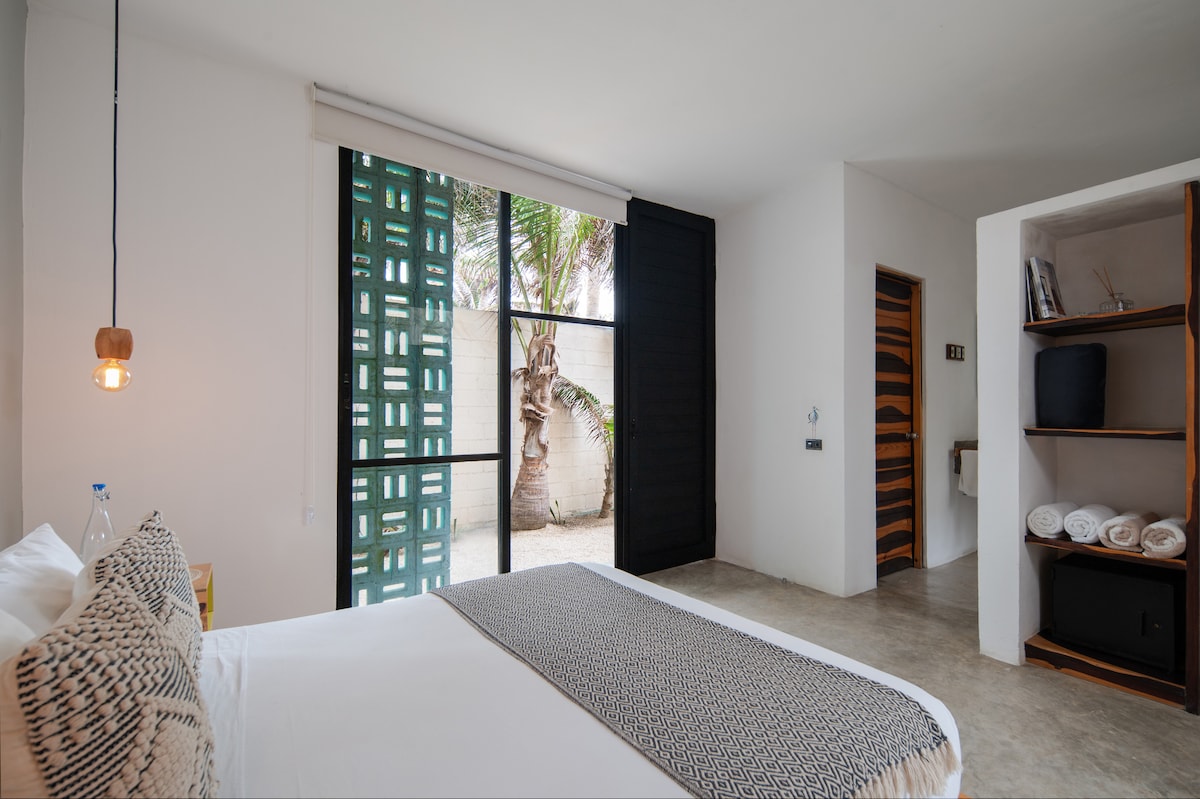 A light-filled bedroom features a spacious bed with patterned pillows, framed by large glass doors that open to a private outdoor area. A modern shelving unit with towels and decor is positioned nearby, complemented by minimalist design elements and a neutral color palette.