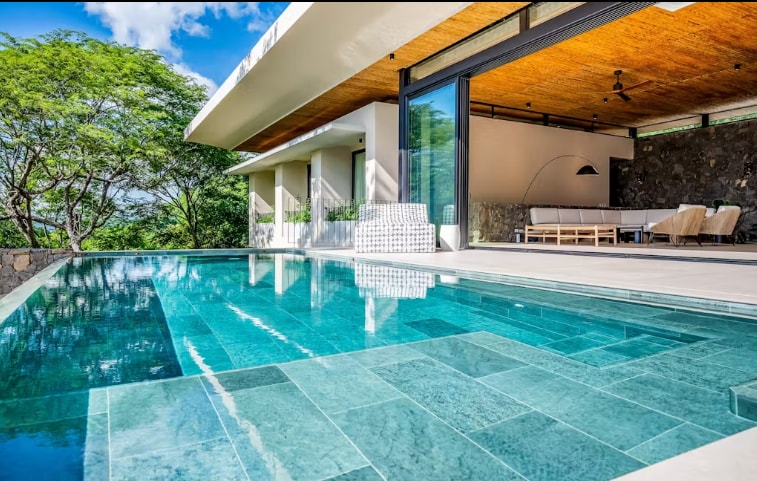 An infinity pool reflects the blue sky and surrounding greenery, seamlessly connecting to an open terrace. The spacious outdoor area is accented by modern lounge furniture, creating an inviting space for relaxation and enjoyment of the tranquil surroundings.