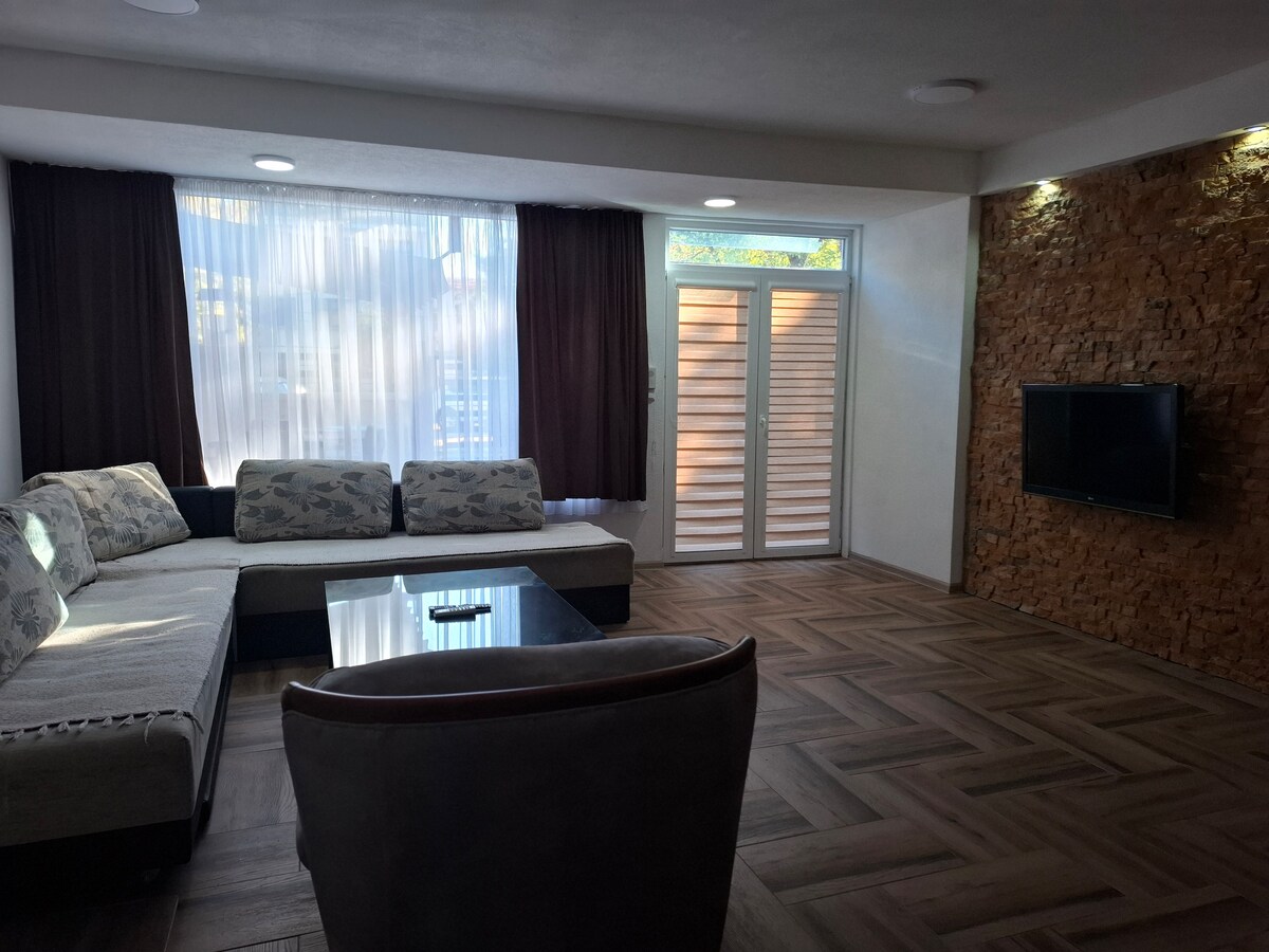 A spacious living area features a large sectional sofa with decorative cushions, complemented by a glass coffee table. Natural light is filtered through sheer curtains, and a flat-screen TV is mounted on the wall beside a textured stone accent. Patio doors lead outside.