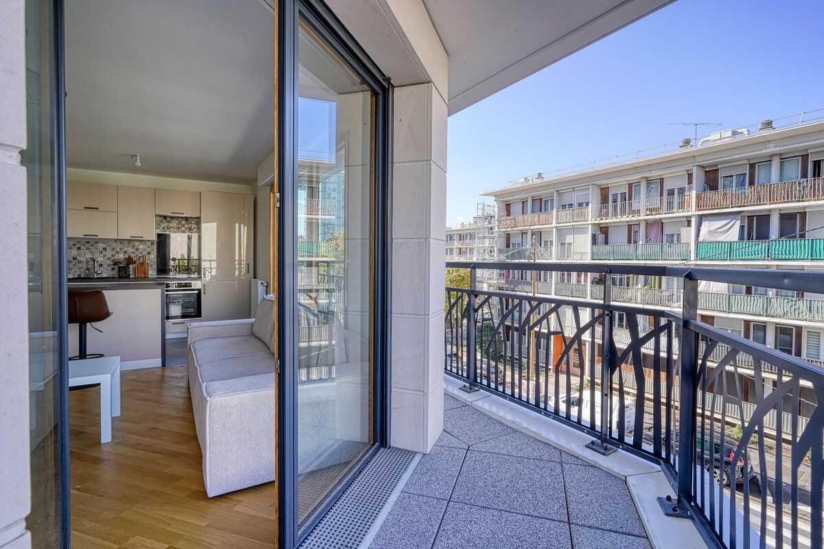 A balcony opens to a view of the surrounding buildings, showcasing a blend of modern architecture. The living area features a light-colored sofa and a compact kitchen visible through large glass doors that invite ample natural light into the space.