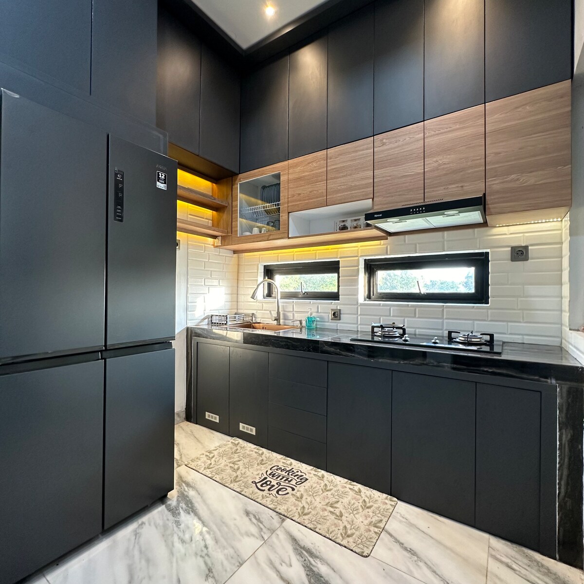 A modern kitchen displays sleek black cabinetry and a contrasting wooden accent. Stainless steel appliances, including a refrigerator and a gas stove, are arranged for functionality. Ample natural light enters through windows, illuminating the white tiled walls and marble flooring.