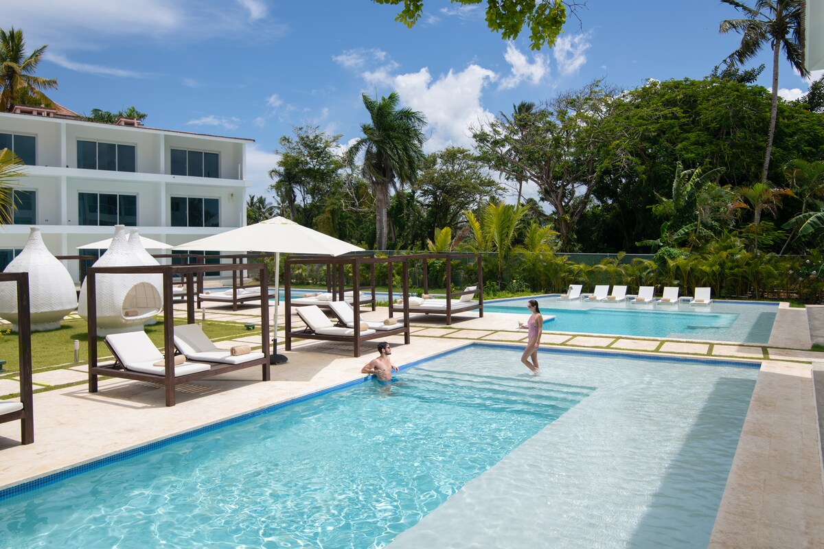 A serene pool area is visible, featuring two swimming pools surrounded by sun loungers and shaded areas. The landscape includes lush greenery and palm trees, contributing to a tropical atmosphere. A clear blue sky complements the inviting water.