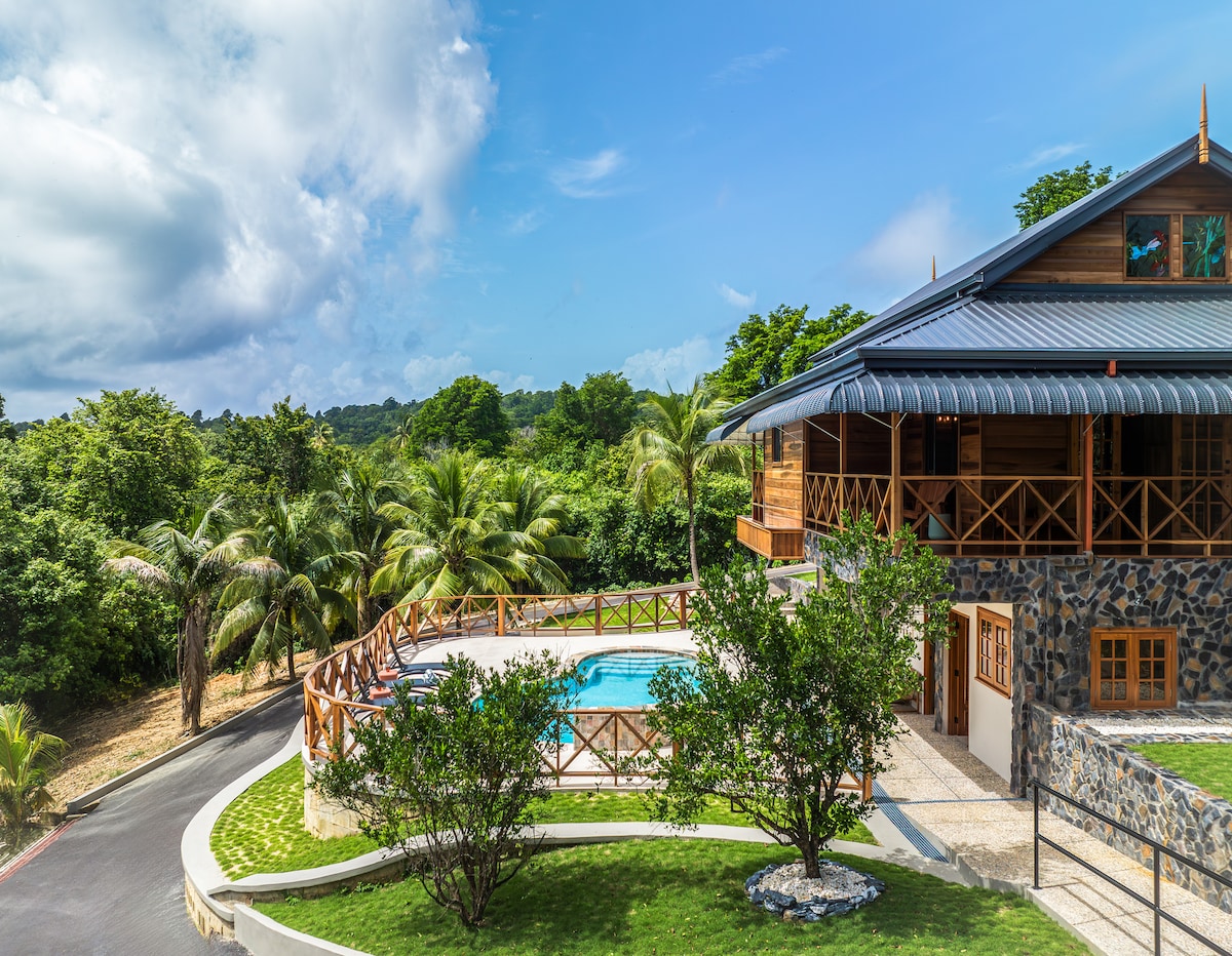 The villa is surrounded by lush greenery, featuring a pool nestled within a fenced outdoor area. A wrap-around porch with wooden railings offers views of the landscape. The two-story structure showcases a blend of natural materials and earthy tones, creating harmony with the environment.